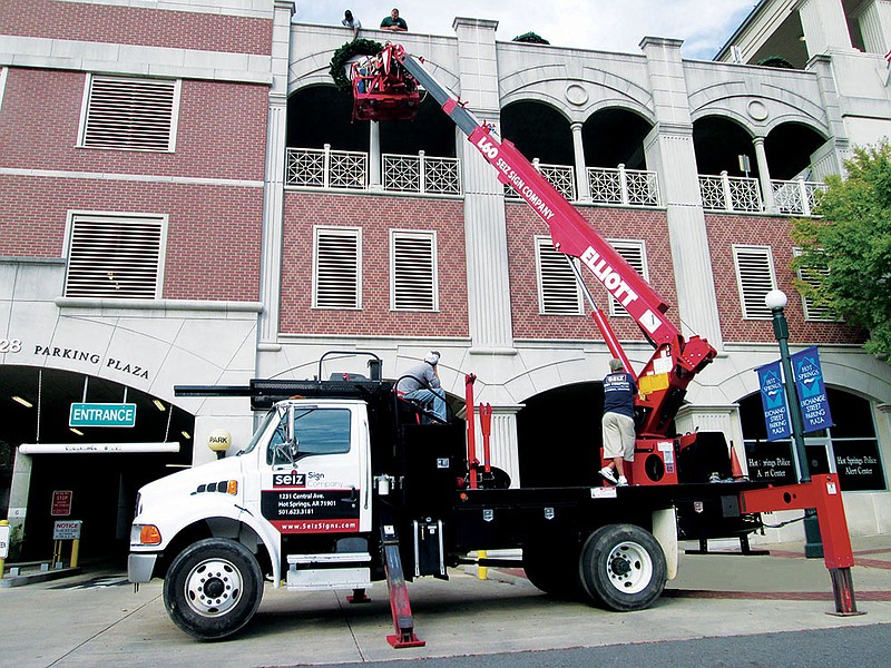 Hot Springs Parks Maintenance Division personnel install a new 6-foot lighted Christmas wreath on the upper facade of the Exchange Street Parking Plaza in downtown Hot Springs. For 11 years, city workers have used bucket trucks from Seiz Signs to place the lights for the holidays.