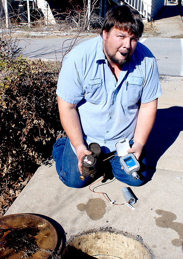 Photo by Dodie Evans Cory Eoff, employee with the Gravette Water Department, holds a plumbing fitting between his teeth as he prepares a new electronically-read water meter at a customer s meter box. He removed the old mechanical meter, which he holds in his hands, and prepares to install the new device which transfers water consumption information directly to City Hall. This can help recognize almost instantly if a customer has a bad leak in the plumbing. Such information, relayed to the customer, can possibly save him many dollars on a water bill. About 150 of the more than 1,000 meters are in the process of being installed throughout the city.