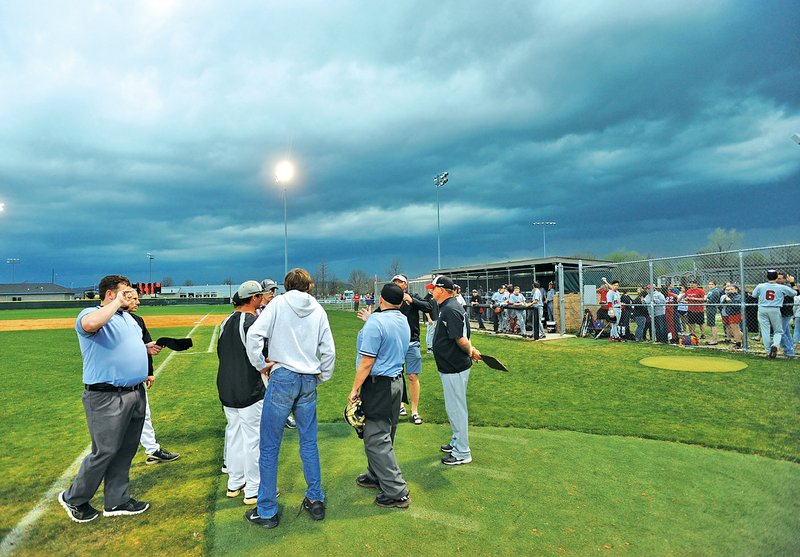 STAFF PHOTO ANDY SHUPE Prairie Grove and Farmington coaches gather Thursday near the plate with umpire Steve Zega to call the game in the seventh inning as storm clouds roll into Prairie Grove.