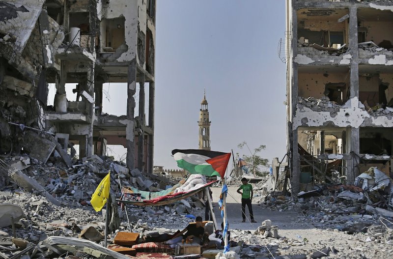 Back dropped by the damaged minaret of the Al-Azba mosque, Palestinians sit under a tent they erected by the rubble of houses, destroyed by Israeli strikes during the war, in the town of Beit Lahiya, northern Gaza Strip, on Tuesday, Aug. 12, 2014. 