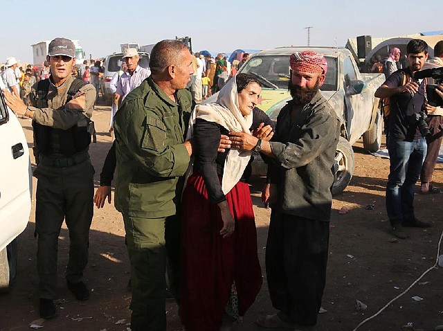 Syrian Kurdish peshmerga fighters help a sick Yazidi woman to a clinic Tuesday inside a Syrian refugee camp where she and other Iraqi refugees have landed after escaping Iraq with the aid of the fi ghters. An Iraqi military helicopter ferrying aid to refugees stranded in northern Iraq crashed, killing the pilot.
