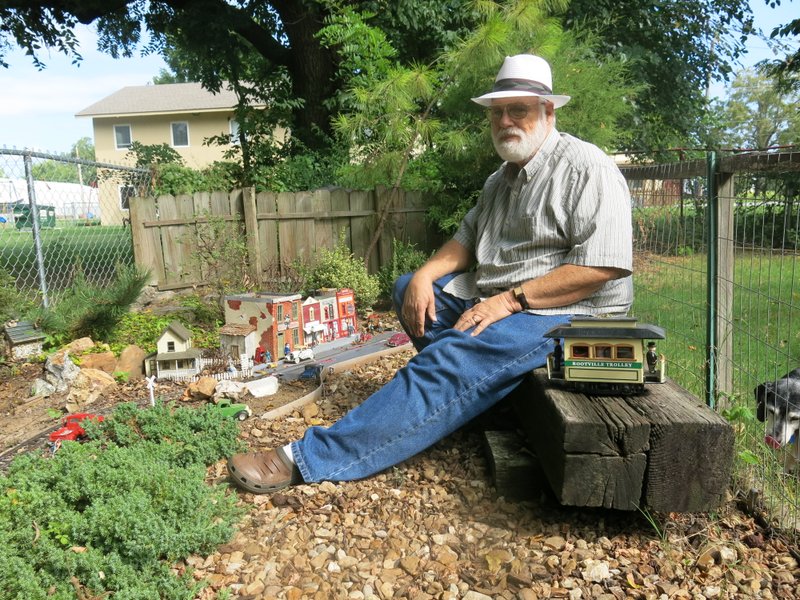 Photo by Susan Holland Jeff Davis relaxes on the viewing bench he built for visitors to his garden railway layout. Sitting beside him on the bench he built from railroad ties is the Rootville Trolley, another of his creations. In the background the tiny town of Rootville can be seen, encircled by tracks from his Spavinaw and Honey Creek Railway.