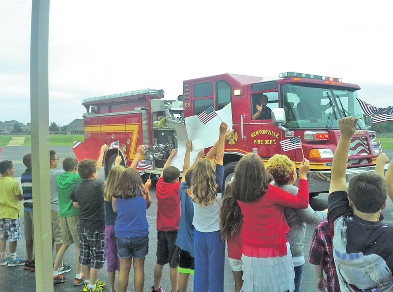STAFF PHOTO Dave Perozek Elm Tree Elementary students wave flags and cheer as a Bentonville fire engines circles a lot behind the school during an event held Thursday to honor first-responders. Bentonville police also participated in the parade.