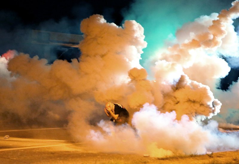 In this Aug. 13, 2014 file photo a protester takes shelter from smoke billowing around during protests against the shooting of 81-year-old Michael Brown in Ferguson, Mo. A federal judge on Thursday, Dec. 11, 2014, granted a temporary restraining order on behalf of protesters involved in the demonstrations that requires police to warn crowds of impending use of tear gas and provide "reasonable" time for people to disperse before tear gas is deployed. The ruling applies only to Missouri.