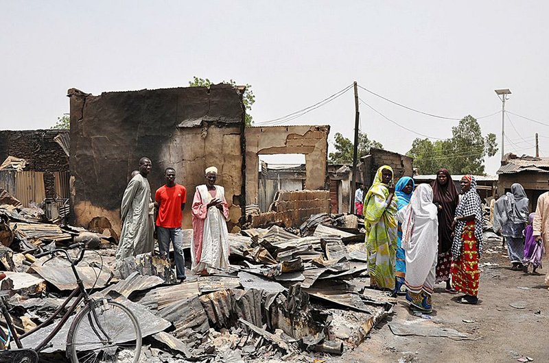 FILE- In this Sunday, May 11, 2014 file photo, people stand outside burnt houses following an attack by Islamic militants in Gambaru, Nigeria. Thousands of members of Nigeria’s home-grown Islamic extremist Boko Haram group strike across the border in Cameroon, with coordinated attacks on border towns, a troop convoy and a major barracks. Further north, Boko Haram employs recruits from Chad to enforce its control in northeastern Nigerian towns and cities. In Niger, the government has declared a “humanitarian crisis” and appealed for international aid to help tens of thousands of Nigerian refugees driven from their homes by the insurgency. (AP Photo/Jossy Ola, File)