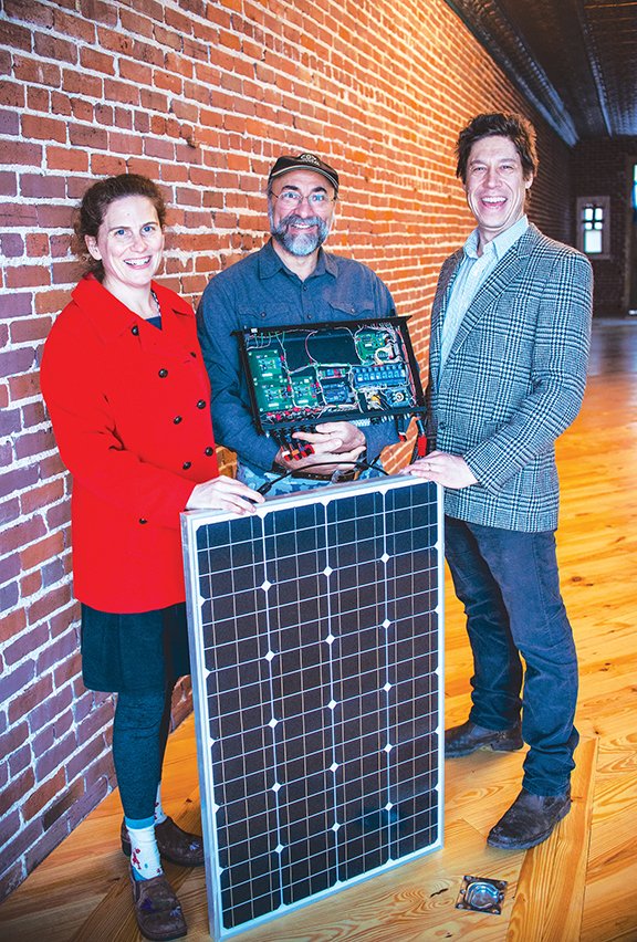 Cheryl Roorda, left, radio station “mom”; Bob Nagy, an engineer holding the telemetry and remote-control power supply or “brain” of the system that runs the future radio station on DC current; and Zac Smith, station manager, stand with a solar panel in the space along Ouachita Avenue that will become the station.