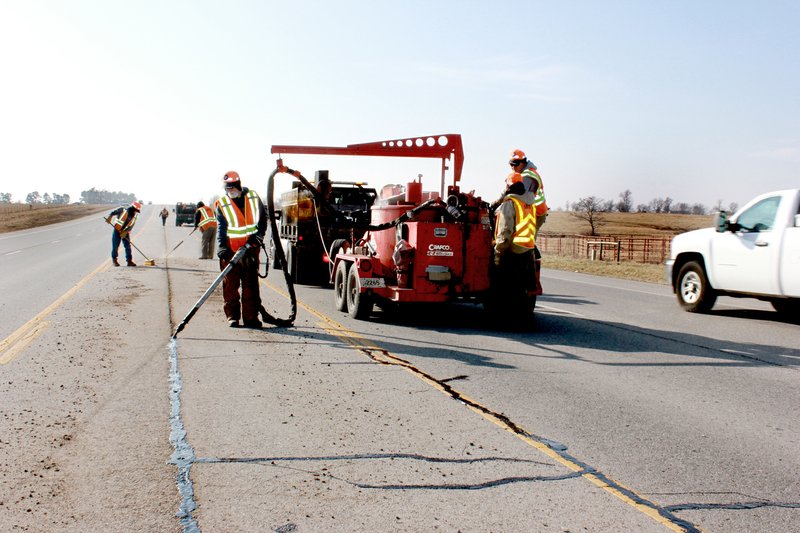 Tina Parker/Siloam Sunday Workers from the Arkansas Highway Transportation Department use compressed air to blast gravel and debris from cracks in Arkansas Highway 59 near Cheri Whitlock Drive on Friday. Once the cracks are cleared, a tar-like substance is pumped into the crack to seal out moisture. The crew has been working its way south from Gentry for the past week.