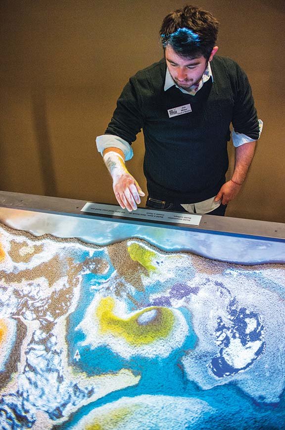 Jim Miller, director of marketing for the Mid-America Science Museum in Hot Springs, holds his hand over an interactive display that allows a person to change the terrain by moving land and water shown on the display, which is intended to illustrate how water changes course when the land is changed.