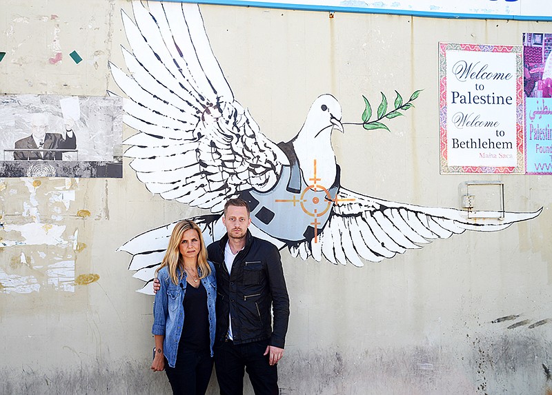 The Associated Press COOKING MEETS CONFLICT: In this image provided by the Travel Channel, journalist Mariana Van Zeller, left, and chef Michael Voltaggio, hosts of Travel Channel's "Breaking Borders," pose in front of an art piece showing a dove in a bulletproof vest by graffiti artist Banksy in Bethlehem, Palestine. The show is a travelogue, cooking show and dining-table summit, premiering today.