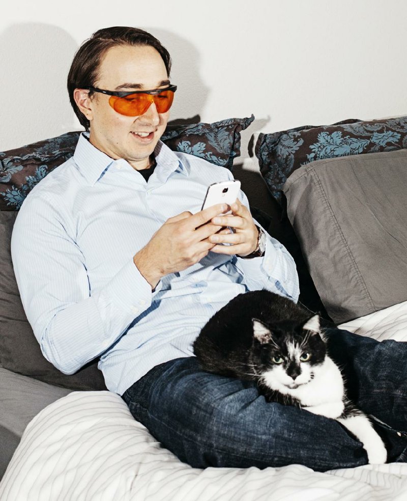 Matt Nicoletti checks his phone on his bed with his cat, Bruce, while wearing orange glasses at his home in Denver, April 1, 2015. Glasses designed to make it easier to fall asleep, by blocking blue light from electronic screens, are growing popular, but are largely untested. (Benjamin Rasmussen/The New York Times)