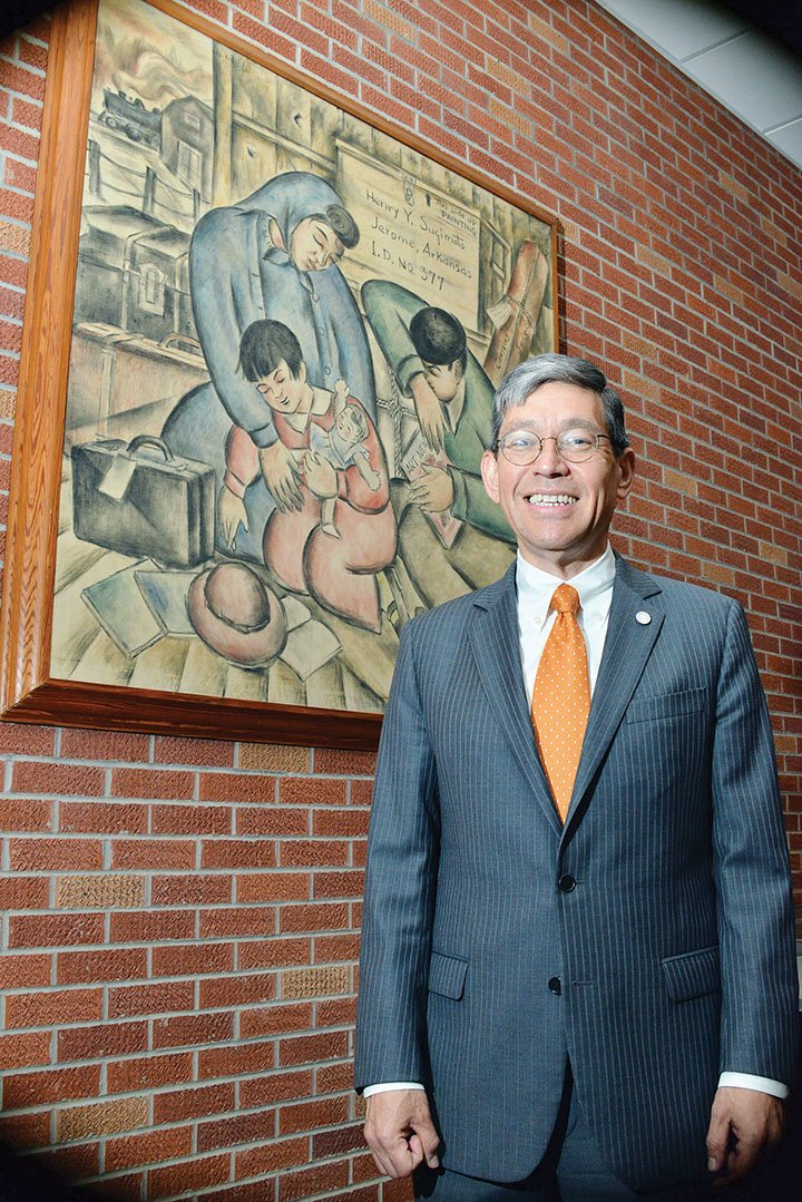 Hendrix College President Bill Tsutsui stands in front of a painting by Henry Sugimoto, an artist who was interned at Jerome Relocation Camp during World War II. Two Hendrix College professors, including Louis Freund, visited the camp and arranged a monthlong exhibit of Sugimoto’s work in 1944. After the exhibit, the college bought Arrival at Camp Jerome, which hangs in the Mills Center on campus. The painting will be commemorated Friday during inauguration events for Tsutsui, a Japanese-American and the 11th president of Hendrix, and Sugimoto’s daughter, depicted in the painting, will attend.