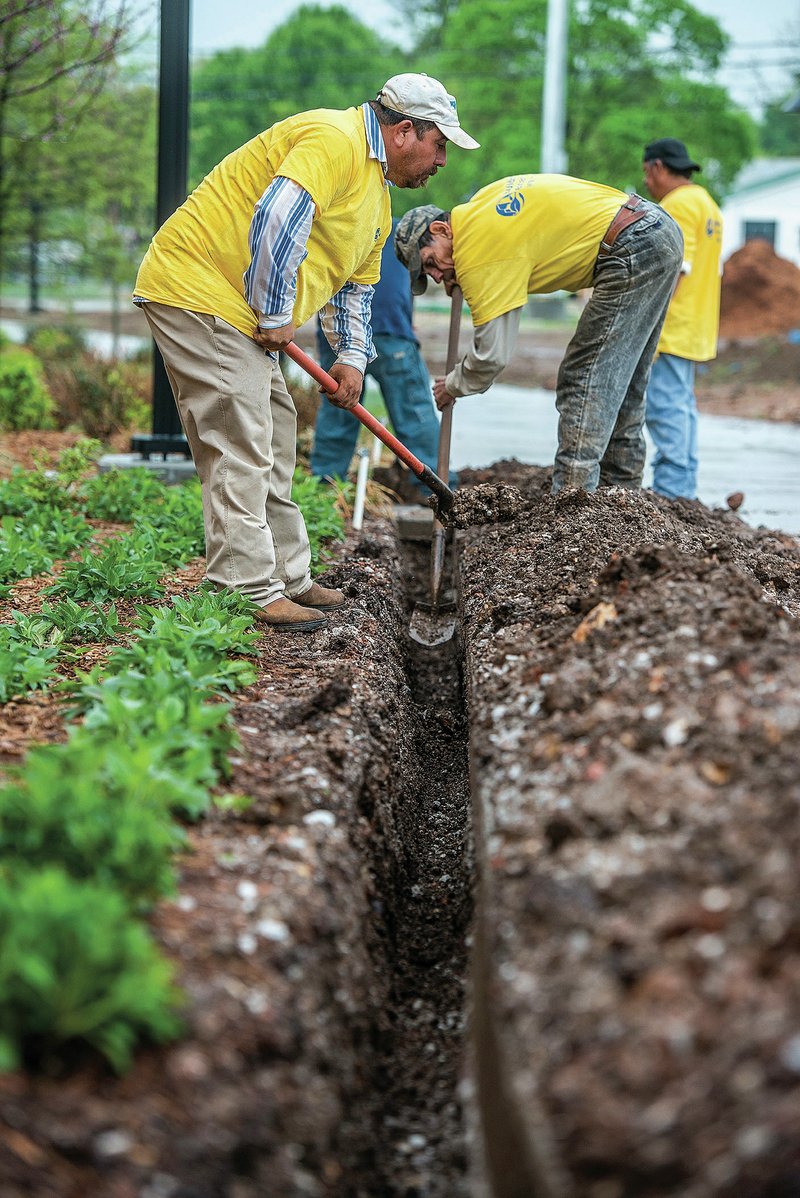 Pedro Rico (left) and Gabriel Mendoza, both with Building Exteriors, dig a trench Friday at Shiloh Square in downtown Springdale. The crew was installing water and drain lines for drinking fountains for use at the square and the Razorback Greenway