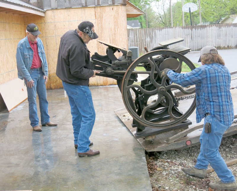 Photo by Susan Holland Gravette Museum commission chairman John Mitchael, left, watches with interest as Gary Phillips and Dale Harrington, owner of D&amp;C Wrecker Service, start to set an old hand press onto the concrete slab on the west side of the museum. The press, which was used for hand printing for years at the former Gravette News Herald office, was donated to the museum by Dodie and Louise Evans and will be housed in the new museum annex when it is completed.