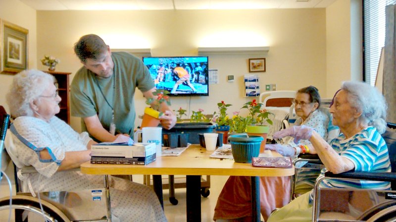 Photo by Debbie Cousineau Will Denver, nurse aide at Ozarks Community Hospital in Gravette, helped swingbed unit patients Lura Kemp, Norma Schmitz and Lorraine Hall plant flowers in pots they had decorated in celebration of Earth Day April 22. It was too cold and windy to go outside on Wednesday, so the ladies decided to do their planting indoors. Pots were filled with petunias, salvia and pansies; and patients will have a pot to take home when they are dismissed from OCH. They also planted extra pots to take to the rooms of patients who were not able to participate. Case manager Debbie Cousineau says they are looking forward to nicer weather when they will go outside and plant more.