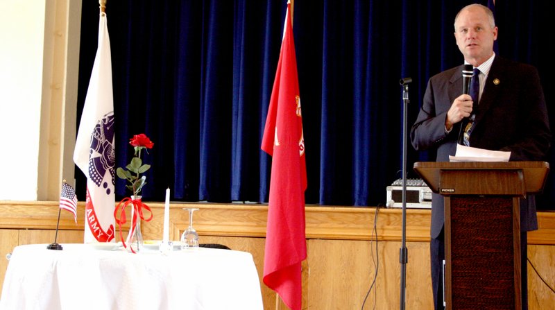 Landon Reeves / Herald Leader Sen. Jim Hendren (R-Gravette) is speaking to the crowd about the origins of Memorial Day and the cost of war. Next to him is a small table for one with a red rose, some salt, a lime wedge, and an inverted glass on a white cloth with an empty seat. Each item is a different symbol that in some way honors or recognizes the soldiers, sailors, airmen, and Marines who died in combat or training, or became a prisoner of war or were missing in action.