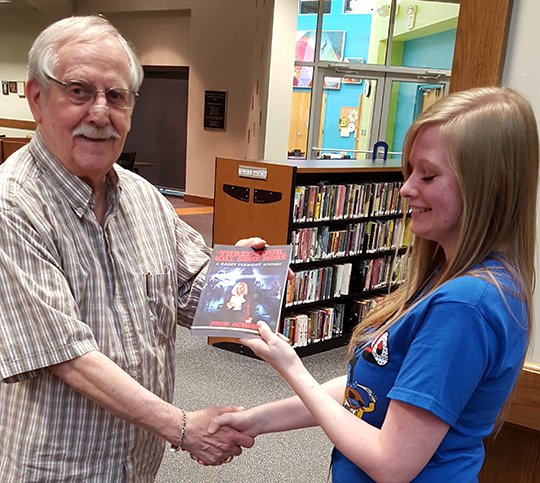 Submitted photo SLEUTH STORY: Area author John Achor, left, presents a copy of his latest mystery novel to Garland County Library staff member Alexis Tomlin. The book is his second in the series.