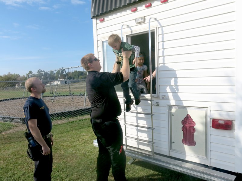 Photo by Susan Holland Gravette firefighter/EMT Wesley Gale helped Glenn Duffy Elementary student Jayden Alexander as he exited the fire prevention safety house last Wednesday at the school. His fellow firefighter, Spencer Gillming, looked on. Students experienced a simulated fire in the &#8220;smoke house&#8221; and were taught how to safely react if they were in a real fire.