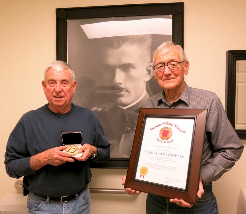 Photo by Susan Holland Bob Kelley and John Mitchael, members of the Gravette Historical Museum commission, display the medallion and certificate awarded to Capt. Field Kindley upon his induction into the Arkansas Military Veterans Hall of Fame. They posed in front of a photo of Kindley taken early in his flying career. The honors were brought to Gravette last month after the induction ceremony Nov. 14 in Little Rock.