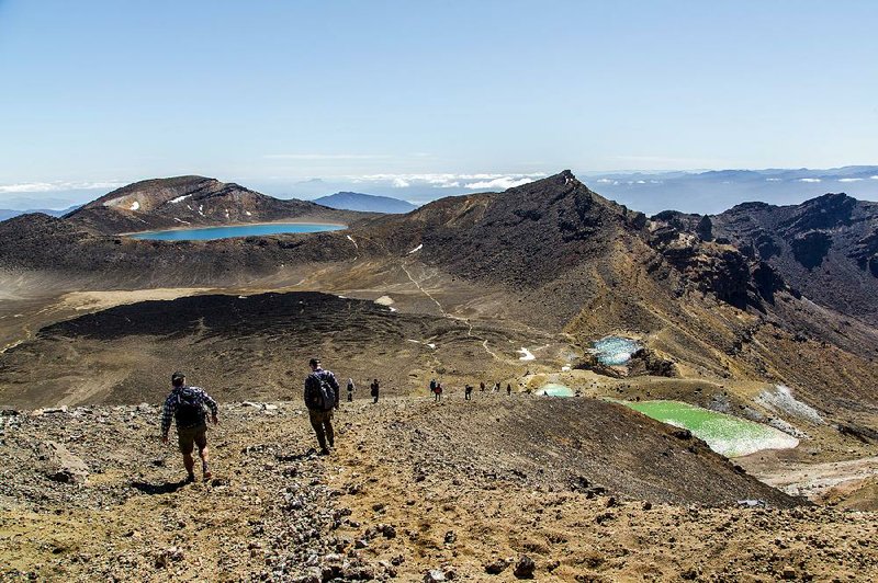 Hikers carefully descend from the Red Crater toward Emerald Lakes (at right) and Blue Lake (far left) on the Alpine Crossing in Tongariro National Park, New Zealand. The largest of the lakes is stained yellow on its edges from the high level of sulphur in the water. 