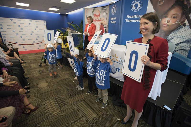 Kathleen McLaughlin (right), president of the Walmart Foundation, and Marcy Doderer (on podium), president and chief executive officer of the Arkansas Children’s Hospital, get some help revealing an $8 million gift from the foundation to the hospital Monday.