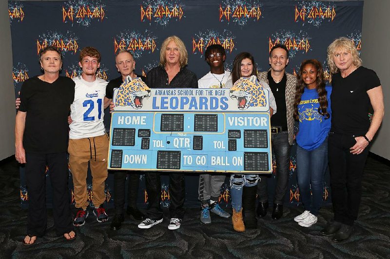 Members of British rock band Def Leppard pose with students from the Arkansas School for the Deaf, whose mascot is the Leopards, before Wednesday night’s concert in North Little Rock. The photo features a replica of the school’s scoreboard. Pictured are (from left) drummer Rick Allen, student Stephen Cathcart, guitarist Phil Collen, lead singer Joe Elliott, students Henry James and Alex Gossett, guitarist Vivian Campbell, student Jewel Brandon and bassist Rick Savage. 