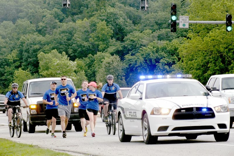 Conor Woody/The Weekly Vista With the Special Olympics Summer Games torch in hand, a group of runners head south on U.S. Highway 71 early Monday morning on the first leg of the torch&#8217;s journey to Searcy. Shown are, from left, Cpl. J.D. Jordan (on bicycle), officer V.J. Wilson, officer Ross Conn, officer Bobby Warren, Bella Vista communications Director Cassi Lapp and officer Reid Hudgens. The group carried the torch to Peach Orchard Road, where they passed the torch to the Bentonville Police Department. It will be passed from department to department until it reaches Searcy, where the Special Olympics Summer Games will take place starting on May 26.