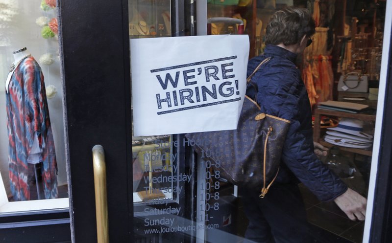 In this Wednesday, May 18, 2016, file photo, a woman passes a "We're Hiring!" sign while entering a clothing store in the Downtown Crossing of Boston. 