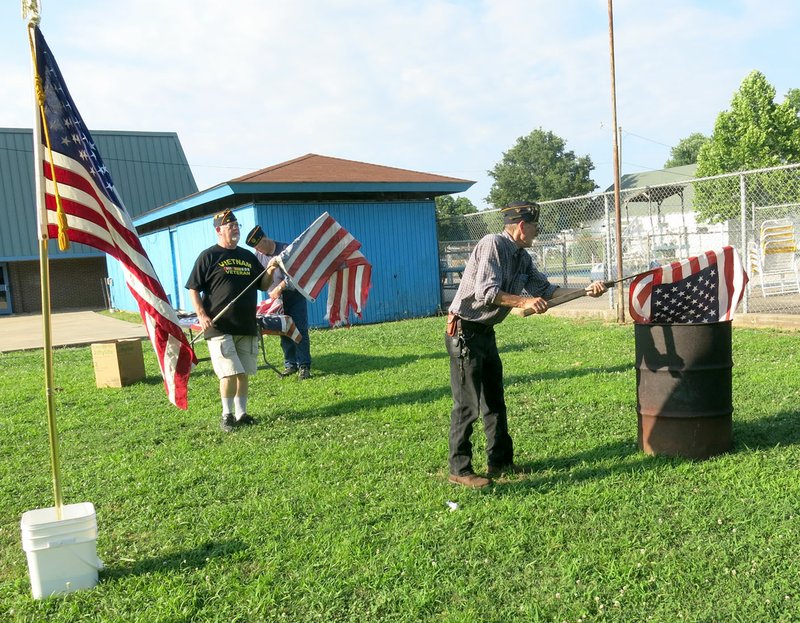 Photo by Susan Holland Dave Malczynski, commander of John E. Tracy American Legion post in Gravette, placed an American flag in the burn barrel as Al Blair, post finance officer, waited to bring another flag to the fire. Mark Russow, second vice-commander of the post, is visible in the background preparing flags. Post members held their annual flag retirement ceremony on Flag Day, Tuesday, June 14, on the Civic Center grounds, followed by a picnic meal.