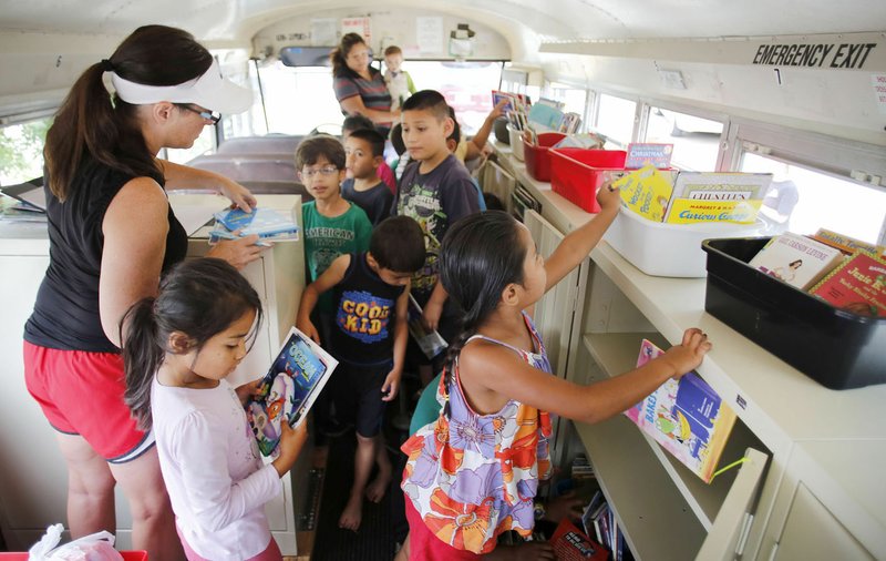 Liesle Tallman (left), an instructional facilitator at Lee Elementary School, helps children check out books Thursday from the school’s Mobile Library at the Holcombe Street Apartments in Springdale. The library recently outfitted a bus and travels through areas of Springdale checking out books to children and distributing snack packs and fresh vegetables when available. 