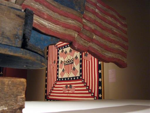 In this June 30, 2016 photo, a patriotic quilt is framed by a flag trailing “Uncle Sam on a Bicycle Whirligig” at the exhibit “American Made: Treasures from the American Folk Art Museum” at the Crystal Bridges Museum of American Art in Bentonville.