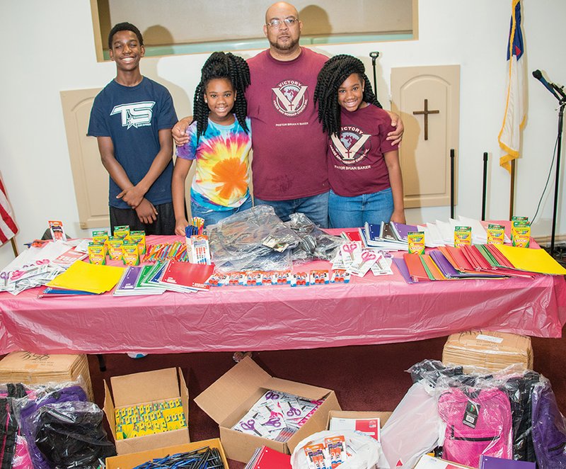 Caleb Campbell, left, 13, Alayna Campbell, second from left, 10, and Alyssa Campbell, right, 10, stand with Victory Praise & Worship pastor Brian K. Baker as they show some of the school supplies the church will give away during its Back-to-School Bash on Saturday. The church will donate 500 mesh backpacks full of crayons, paper, glue and more.
