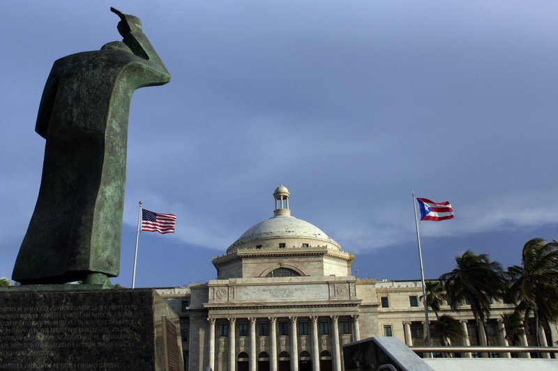 FILE - In this July 29, 2015 file photo, a bronze statue of San Juan Bautista stands in front of Puerto Rico's capitol flanked by U.S. and Puerto Rican flags, in San Juan. 