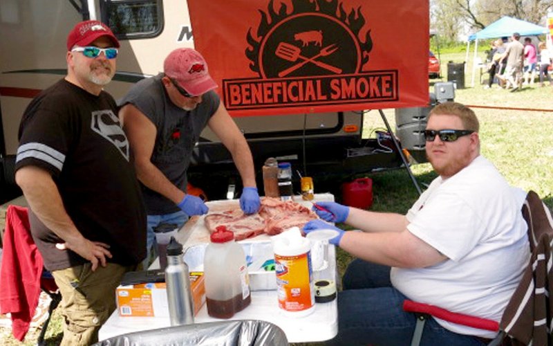 Bobby Cunningham (left) and Matt Rhodes (right), members of the Beneficial Smoke barbecue team from Pea Ridge, found time to pose for the camera while their teammate Brian Hill trimmed fat from the brisket they were preparing to smoke for the Smoke on the Border competition. The Beneficial Smoke team placed 10th in the pork division of the KCBS-sanctioned contest and said they were enjoying the event, their first contest of the year.