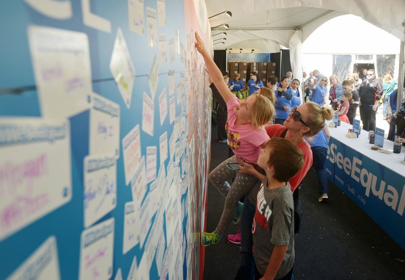 Carrie Schweer of Bentonville lifts her daughter Ellie Schweer, 8, as son Matthew Schweer, 12, looks on Thursday, while adding their stickers to the “We See Equal” wall in the Procter & Gamble booth in the Sponsor Village at Compton Gardens during the Bentonville Film Festival. The family was visiting the festival to celebrate Matthew’s birthday.
