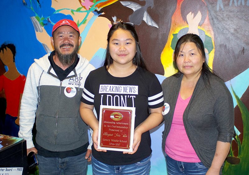 Photo by Randy Moll Julia Lee, an eighth-grade student at Gentry Middle School (center), was awarded a plaque for outstanding achievement in art craftmanship on Friday (May 5, 2017). She is pictured in front of a mural encouraging reading which she is painting on a wall in the school&#8217;s library. With her are her parents, Bee Xiong and Kamashi Lee.