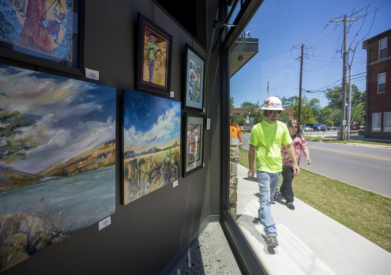 Pedestrians walk along the sidewalk with artwork by Carol Hart displayed May 25 in the window of First National Bank on S.W. A Street in downtown Bentonville.