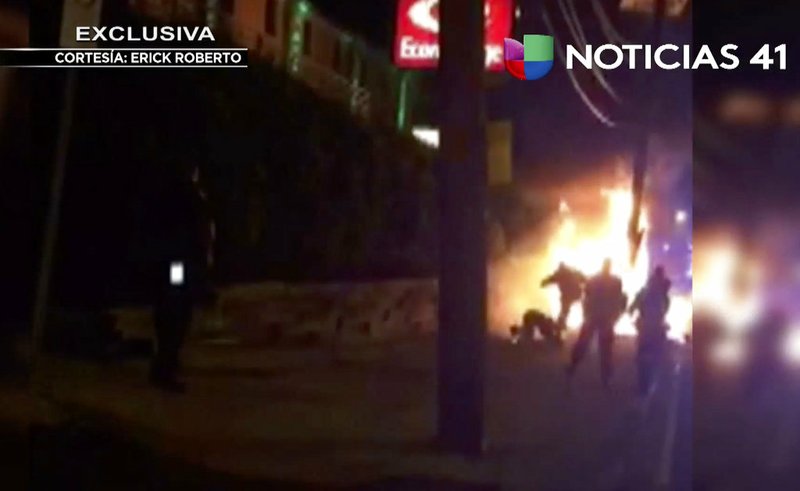 This image from video shot June 4, 2017, shows a bystander being kicked by police officers as he lies on the ground near flames after an automobile chase that ended in a fiery crash in Jersey City, N.Y.