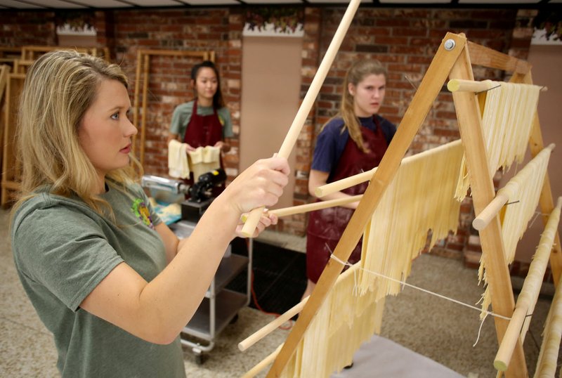 NWA Democrat-Gazette/DAVID GOTTSCHALK Ashley Wiedenhoeft, left, rearranges the dowels on a rack to continue to hang fresh pasta as Hannah Pianalto, 14, and Mary Kidd, 15, work in the background at St. Joseph Parish Hall in Tontitown. More than 3,000 pounds of pasta noodles have been prepared for the 119th Tontitown Grape Festival, which begins Tuesday.