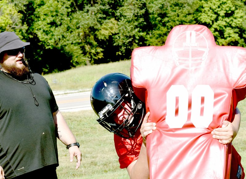 Photo by Rick Peck Line coach Chris Kane looks on as Will Gordon hits a blocking sled during the first day of McDonald County High School&#8217;s fall sports practices on Monday. The Mustangs travel to Reeds Spring on Aug. 11 for a four-team scrimmage before opening the season at Salem on Aug. 18. All games begin at 7 p.m.