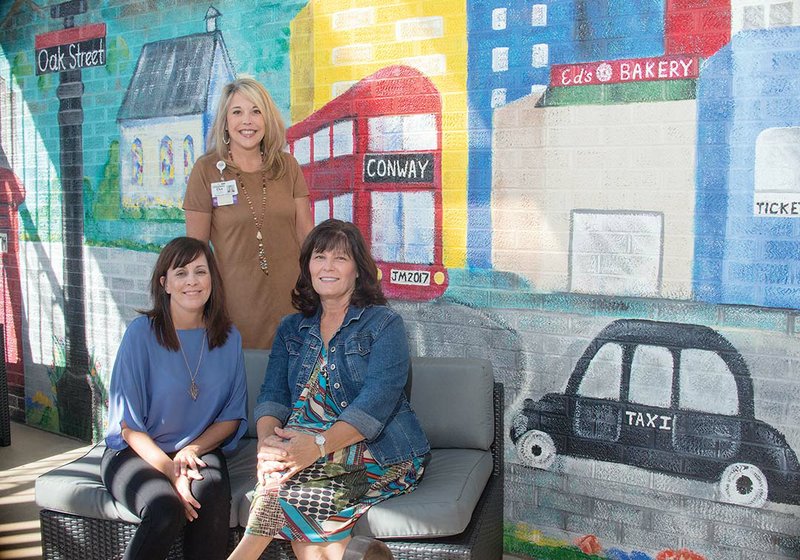 Donna Beshears, left, activity therapist for the Conway Regional Senior Behavioral Health Unit, sits with artist Jan Mallett of Conway in front of the mural Mallett painted on the unit’s patio. Standing is Dot Welch, associate director of the Conway Regional Health Foundation, which funded the mural project. The idea of the mural was to help calm patients with dementia, as well as beautify the area.