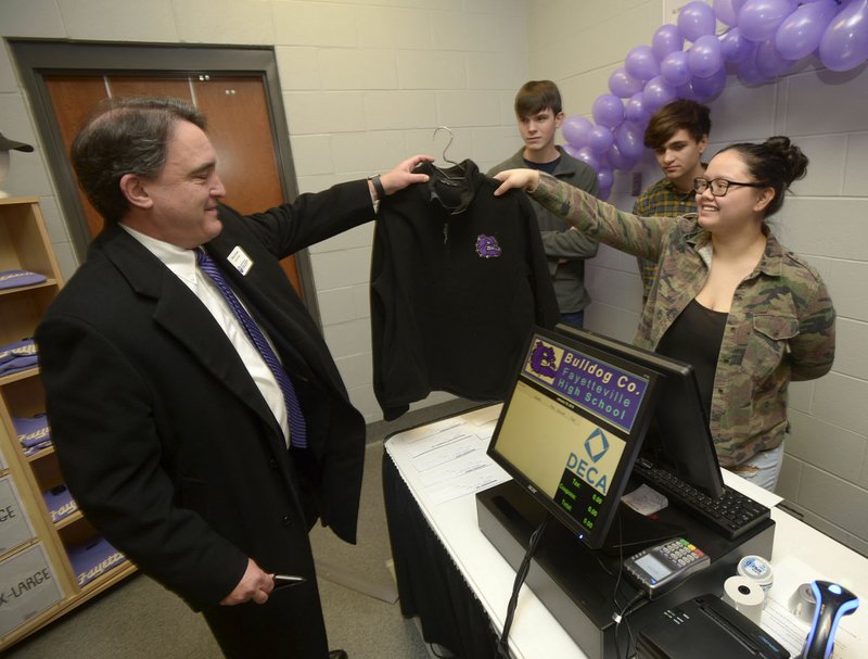 Chris Lawson (left), general counsel for Fayetteville Public Schools, hands over a fleece pullover to Kaly Atienza (right), a junior at Fayetteville High School, as Lawson makes the first official purchase Thursday while Skyler Vaughn (center) and Sam Meullenet look on during a grand opening for Bulldog Co., Fayetteville High School’s school store, at the school. The store serves as a project of the Small Business Operations class at the school, giving students experience in operating a business.