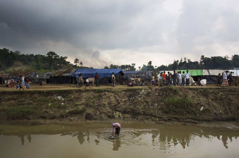 FILE- In this Sept. 4, 2017 file photo, a Rohingya woman living in no man's land washes a pot from a stream near Cox's Bazar's Tumbru area, while smoke rises from fires across the border in Myanmar.  (AP Photo/Bernat Armangue, File)