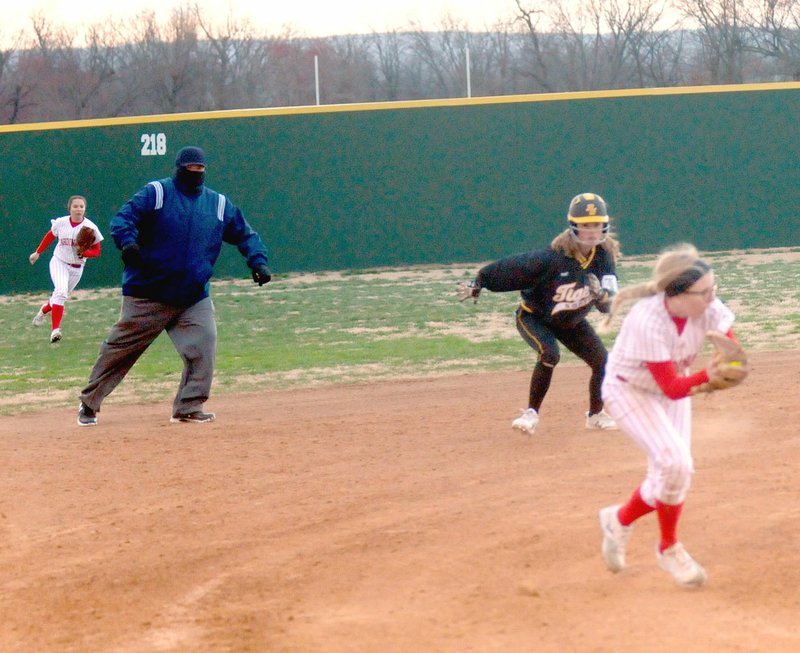 MARK HUMPHREY ENTERPRISE-LEADER Farmington sophomore Paige Anderson scoops up a grounder in front of third while a Prairie Grove base-runner prepares to break for third behind her. The field umpire's garb was indicative of the high winds and cold weather in which the game was played. Farmington won 20-4 by run-rule in three innings on Tuesday, March 6.