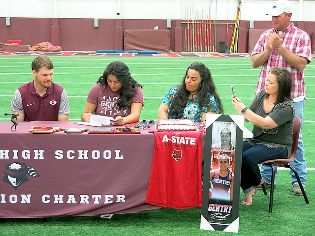 With assistant track coach Ethan Ramsey (left) looking on, Chastery Fuamatu, a Gentry senior and holder of state titles, signs her letter of intent to attend Arkansas State University, and throw the shot and discus there, during ceremonies at Gentry High School on Thursday, April 12, 2018. Her mother Alexandria Fuamatu and family sponsors Ashley and Holly Hays watch and applaud.