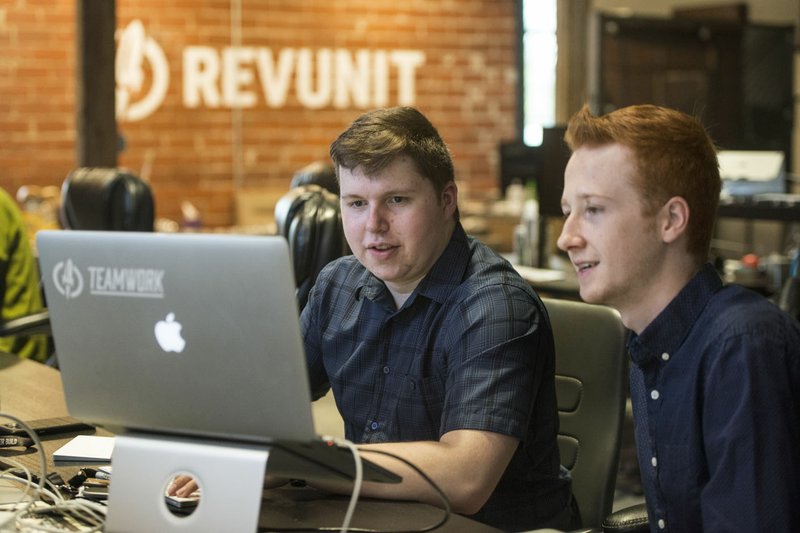 Michael Sutera (left), a senior in the Ignite program at West High School, gets help Thursday from Hayden Holzhauser, a junior developer at RevUnit and a 2016 graduate of the Ignite program at Bentonville High School at RevUnit in Bentonville. Sutera, in the Ignite program’s information technology discipline, is interning at the business this school year. 