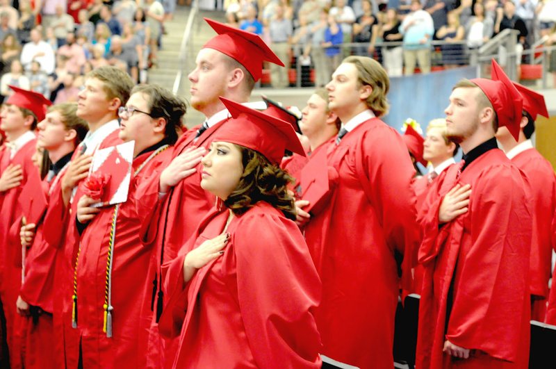 MARK HUMPHREY ENTERPRISE-LEADER Farmington graduates salute the Flag of the United States of America while the Crimson Select Ensemble and the Farmington High School band perform the National Anthem during graduation ceremonies on Tuesday, May 15, 2018, at Cardinal Arena.