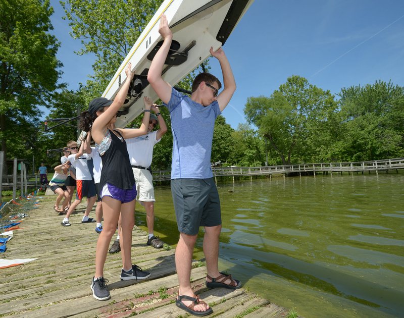 NWA Democrat-Gazette/ANDY SHUPE Campbell Rogerson (center), 17, a junior at Haas Hall Academy, and Ana Estrada, 18, a senior, join others Tuesday in lowering a rowing shell onto the water before participating in a series of informal 1,000-yard sprints to celebrate the graduation of members of the Northwest Arkansas Youth Rowing Club on Lake Fayetteville. Students from Haas Hall Academy and Shiloh Christian School have been learning about rowing from adult members since fall. The club will host novice classes beginning June 30.