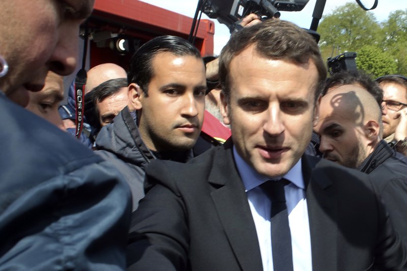 FILE - In this Wednesday April 26, 2017 file photo, Emmanuel Macron, right, is flanked by his bodyguard, Alexandre Benalla, left background, outside the Whirlpool home appliance factory, in Amiens, northern France. Investigators have detained for questioning on Friday, July 20, 2018 one of President Emmanuel Macron's top security aides caught on camera beating a protester in May, a turn of events now evolving into a major political crisis for the president. The presidential Elysee Palace said it is taking steps to fire Alexandre Benalla, who was identified earlier this week by the newspaper Le Monde for beating a young protester during May Day protests while wearing a police helmet (AP Photo/Thibault Camus, File)