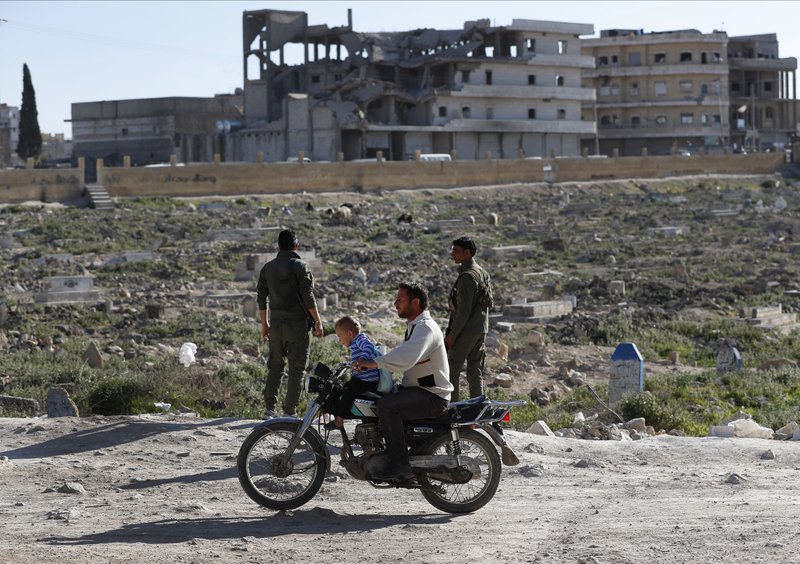  In this March 31, 2018, file photo, a Syrian man with his child rides a motorcycle, as he passes members of the Kurdish internal security forces, at the scene where an explosion hit U.S.-led coalition vehicles, killing an American and a British soldier, in Manbij, north Syria. (AP Photo/Hussein Malla, File )