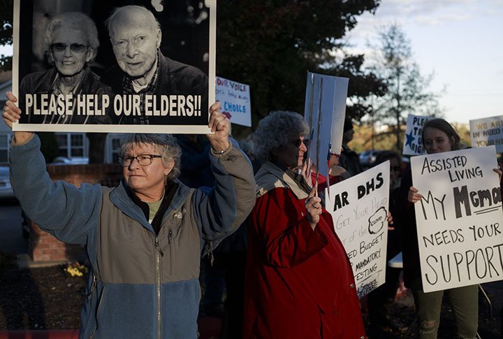Marilyn Knapp of Fayetteville (left) holds a sign showing her parents during a rally Nov. 2 at the Gardens At Osage Terrace in Bentonville. About 70 concerned family members attended a rally to protest the proposed DHS rules concerning provider care, rates and changes in the assisted living homes, which is being pushed to be revised and reviewed in a short period of time. “If this amendment goes through as is, they’ll potentially be displaced and I don’t know where they’re going to end up. They may go to a nursing home or they may go with me,” Knapp said.